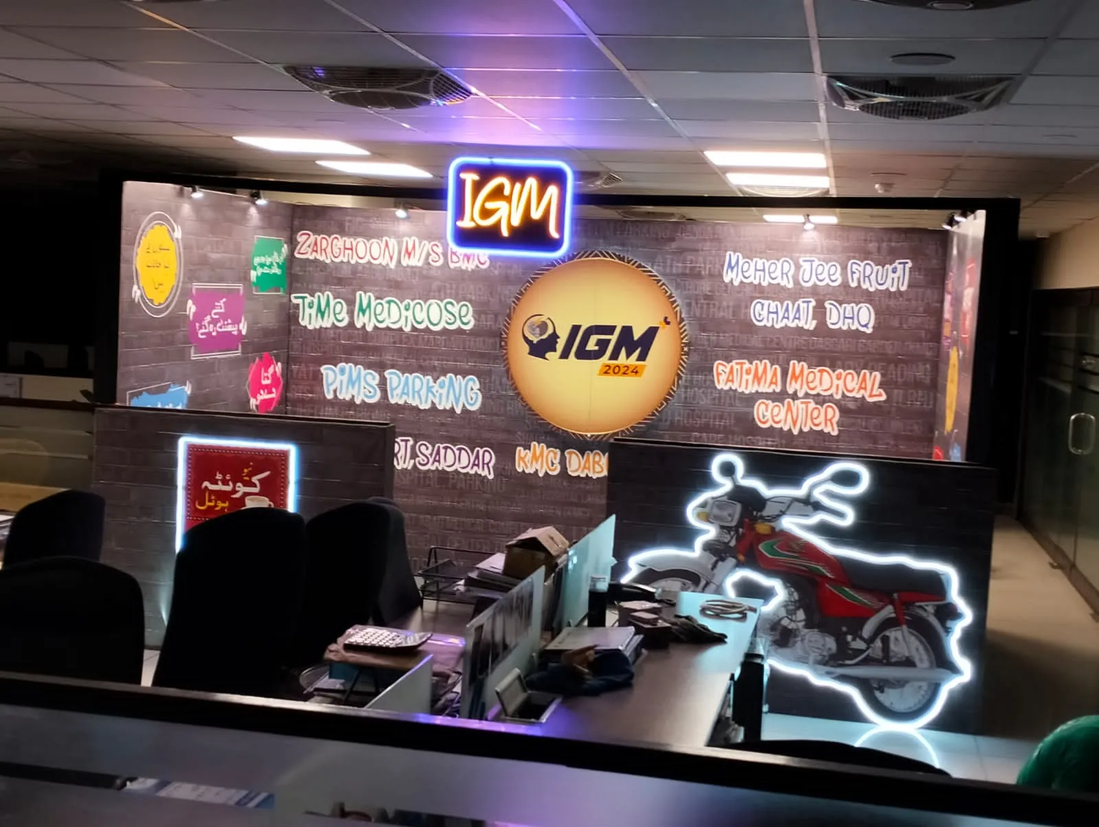 The IGM 2024 exhibition build: a brick-textured wall carrying neon-lit IGM lettering, hand-painted signage panels in the style of Pakistani street art, a large illuminated IGM 2024 disc, and a neon-outlined motorcycle display piece.
