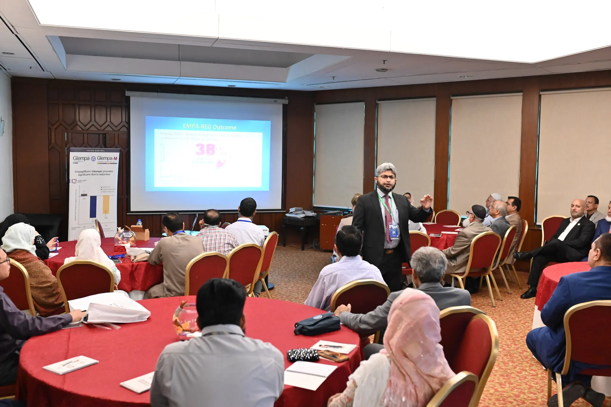 A round-table breakout session at PCDA Symposium 2024, with a speaker addressing seated delegates beside a projection screen and a branded standee.