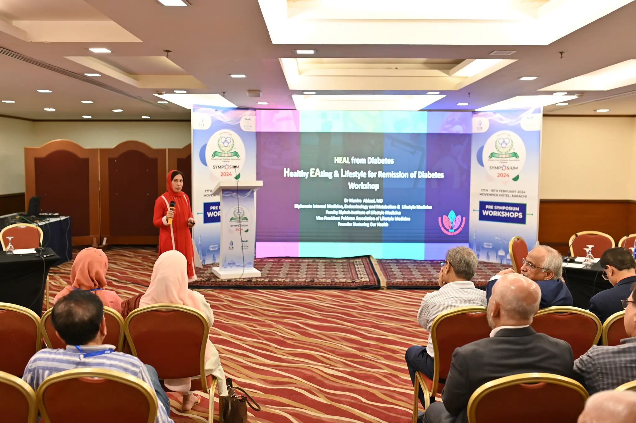 A pre-symposium workshop in progress at PCDA Symposium 2024, with a speaker presenting beside a branded lectern in front of a wide LED wall carrying the session title.