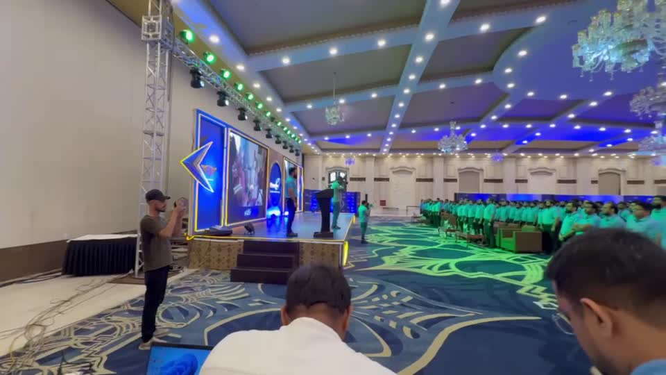 A ballroom set for a large corporate conference: rows of delegates in front of a wide LED stage, lit in blue, with trussing and chandeliers overhead.