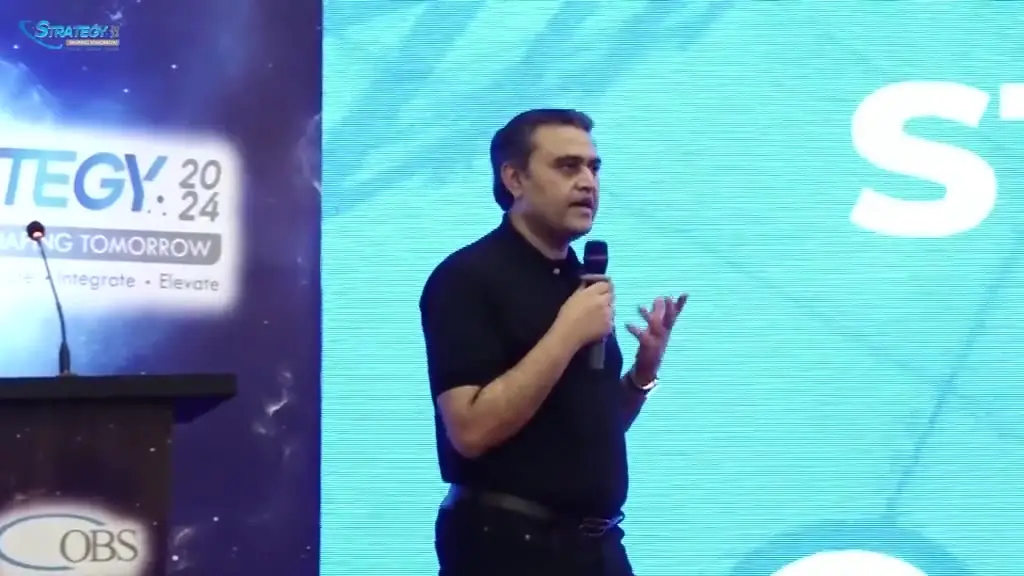 Footage from the Strategy 2024 summit: a speaker presenting at a branded podium in front of an LED backdrop carrying the summit identity, flanked by blue and white balloon columns, with a dressed banquet floor beyond.