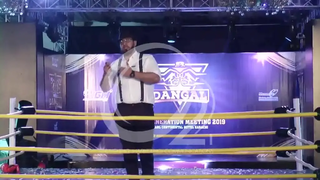 Footage from the 2019 Idea Generation Meeting: a full-size wrestling ring built as the event stage, with yellow ropes, themed arena graphics on the LED wall behind, and speakers presenting from inside the ring under stage lighting.