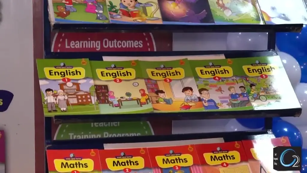 Footage from the Ilm Book Fair and Health Festival: wide convention hall aisles lined with exhibitor book stalls, visitors browsing, and a branded programme stage hosting a health demonstration.